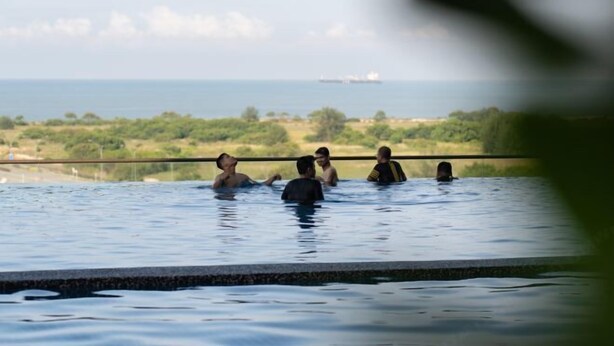 A pool with people swimming, a distant ship, and a view of greenery and hills.