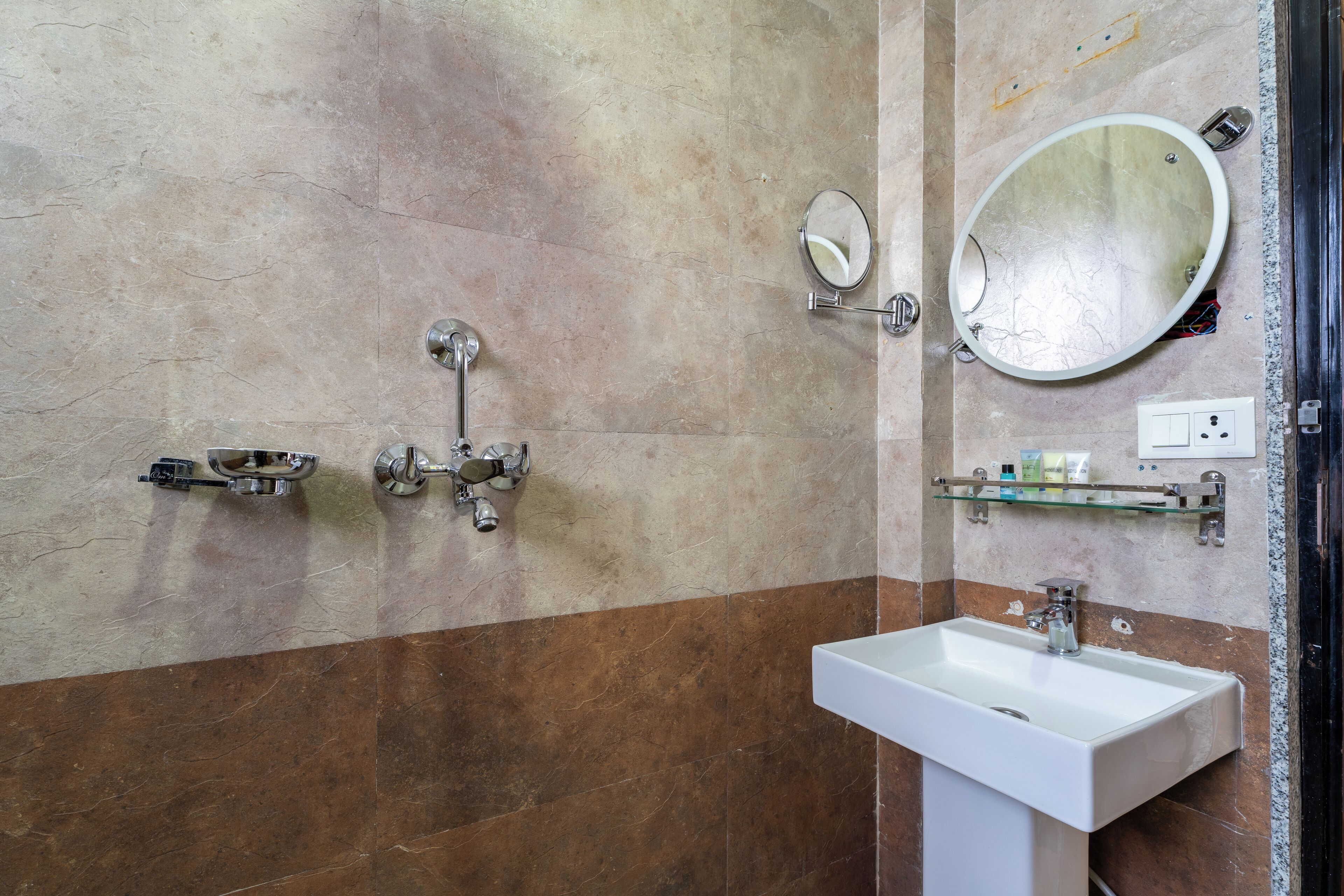 A bathroom with a white sink, two faucets, a mirror, and a glass shelf with toiletries.