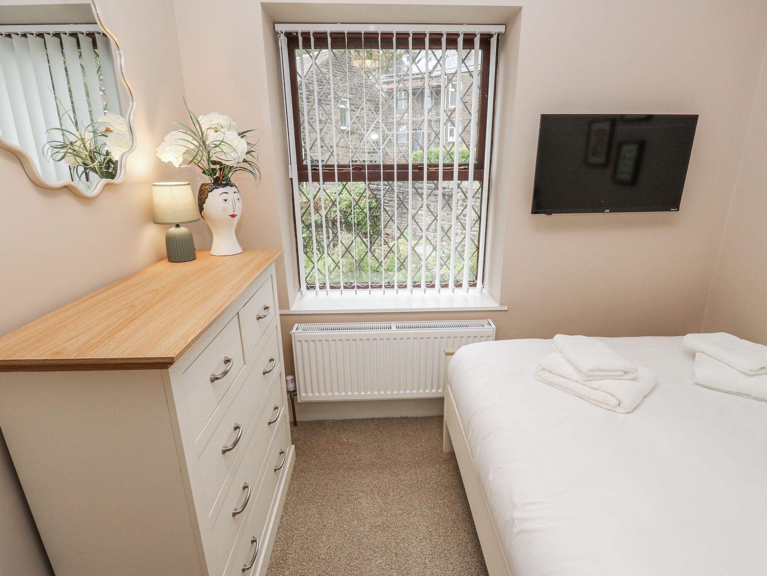 A bedroom with a wooden dresser, a radiator, a bed with white linens, a window with bars, and a wall-mounted TV.