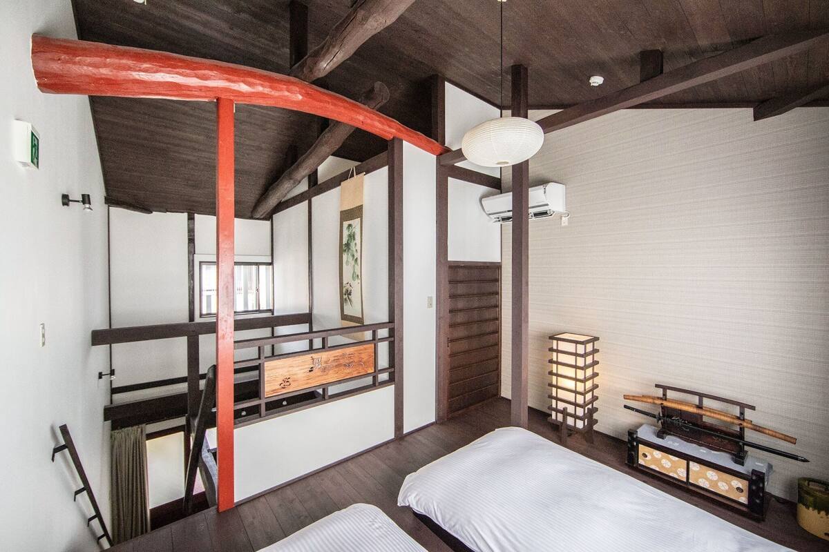 Quarto em estilo japonês tradicional, com teto de madeira, chão de esteira de tatame, cama de madeira baixa, mesa pequena com abajur e porta deslizante para outro cômodo.
