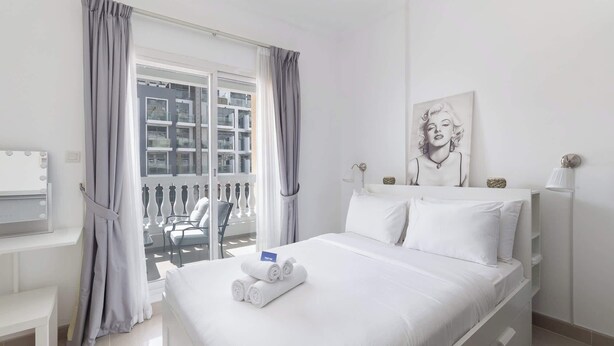 A hotel room with a bed, a desk with a vanity mirror, a balcony with curtains, and a painting on the wall. - JVC 1 bed for 3 People With Swimming Pool (Dubai)