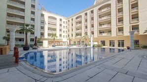 A hotel courtyard with a central swimming pool, multiple balconies, and palm trees. - JVC 1 bed for 3 People With Swimming Pool (Dubai)