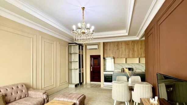 A spacious living room with a sofa, coffee table, and a TV. There is a dining area with chairs and a table. The room has a chandelier and a kitchen in the background. - Apartments in Tashkent City 2 (Tashkent)