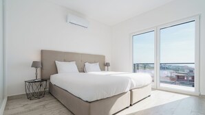 Une chambre spacieuse avec un grand lit, deux tables de chevet et une vue sur le paysage urbain, visible à travers une porte-fenêtre coulissante.