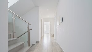 Un couloir moderne avec un escalier à garde-corps en verre, un intérieur épuré aux couleurs blanches et une porte visible au fond.