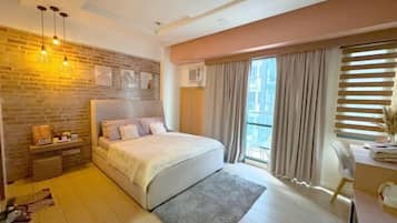 A bedroom with a bed, a desk, a chair, a window with curtains, a brick wall, and a hanging light fixture.