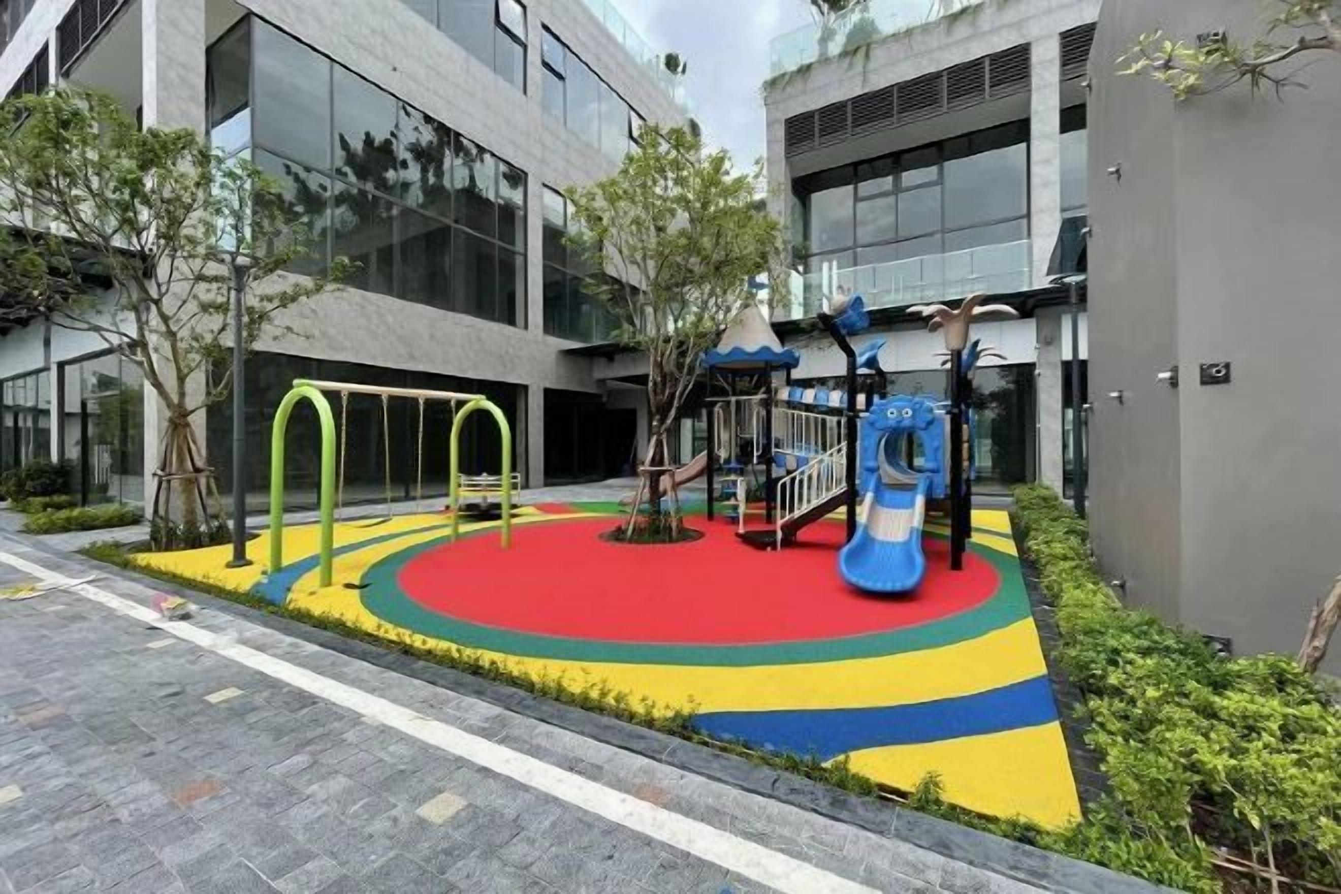 Un'area giochi con altalene e scivoli, circondata da una zona colorata in rosso, giallo e blu, con edifici sullo sfondo.