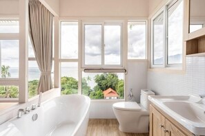 A bathroom with a bathtub, toilet, and sink, large windows with a view of greenery and a house, and a wooden floor. - Grand view @lamaibeach (Koh Samui)