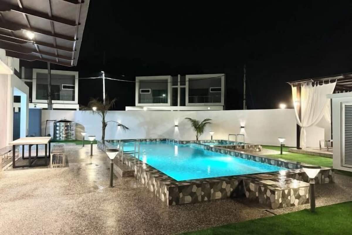 A swimming pool with a stone border, a patio area with a table and chairs, and a building with large windows in the background.
