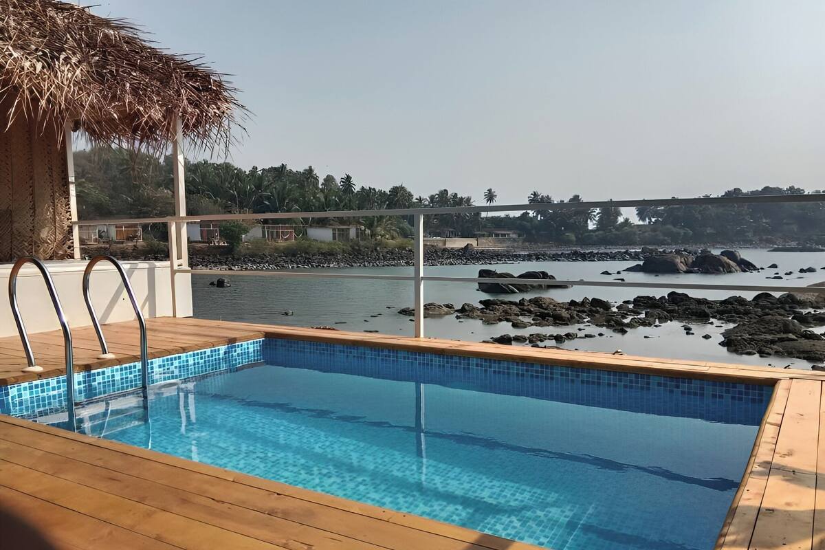 Une piscine dotée d’une terrasse en bois, d’une structure surmontée d’un toit de chaume, et offrant une vue sur une côte rocheuse et des palmiers.