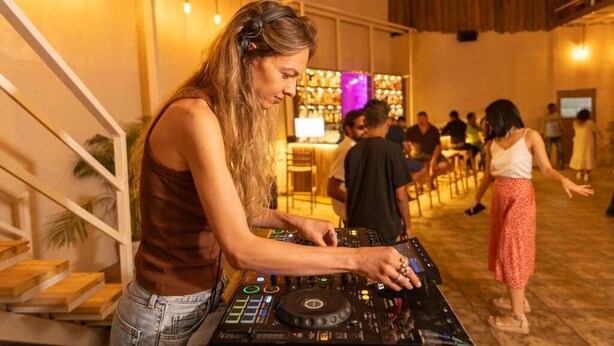 Una persona manejando equipo de DJ en un espacio interior bien iluminado, con una escalera y una zona de bar al fondo.