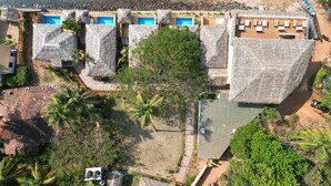 An aerial view of a resort with multiple thatched-roof buildings, a swimming pool, and a beach. - Destiny Goa Beach Resort (Canacona)