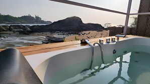 Un área de spa con una bañera grande y una terraza de madera con vistas a una playa rocosa.