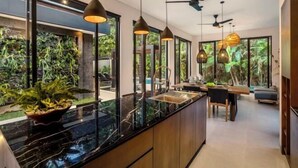 A modern kitchen with a black marble countertop, pendant lighting, and a view of a lush garden through large windows. - Villa Bale Dauh by BaliSuperHost (Bali)