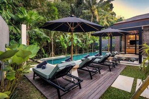 Un área junto a la piscina con sillones y sombrillas, rodeada de exuberante vegetación.