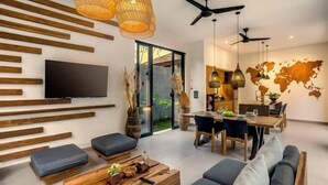 A modern living room with a TV, wooden beams, a sofa, a coffee table, and a dining area with a world map on the wall. - Villa Bale Dauh by BaliSuperHost (Bali)