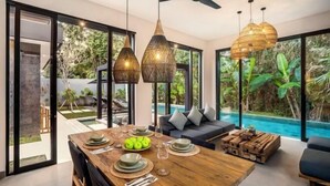 A modern dining area with a wooden table set for four, large windows offering a view of a swimming pool and lush greenery. - Villa Bale Dauh by BaliSuperHost (Bali)