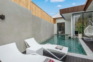 A modern outdoor pool area with lounge chairs, a wooden deck, and a glass door leading to a covered patio. - Villa Serenya Ubud (Bali)