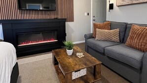 Una sala de estar moderna con chimenea, un televisor de pantalla plana montado en una pared de paneles de madera, un sofá gris con cojines naranjas y una mesa de centro de madera con una planta y un objeto decorativo.