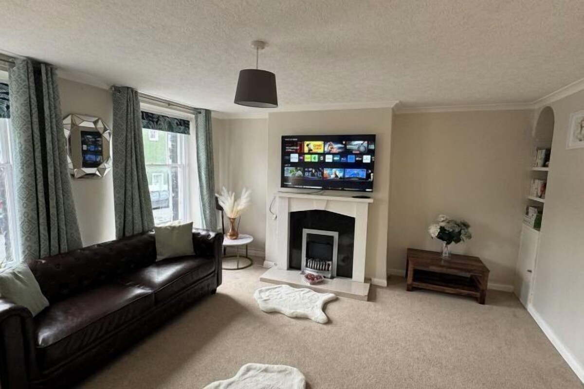 Een woonkamer met een donker leren bank, een open haard, een televisie erboven en een klein houten bijzettafeltje met een vaas.