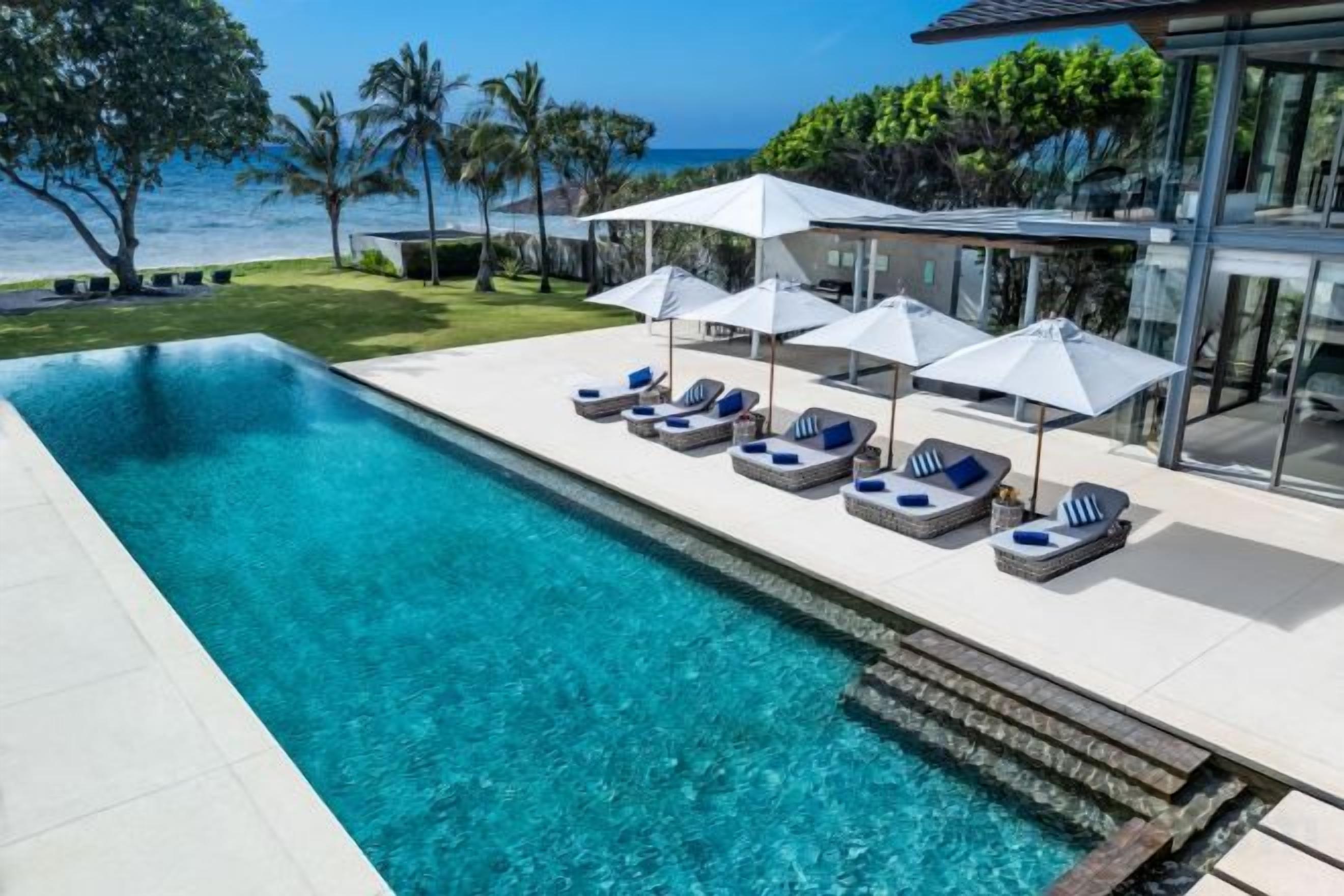 Une piscine dotée de chaises longues et de parasols, une vue dégagée sur l’océan et un bâtiment moderne avec de grandes fenêtres.