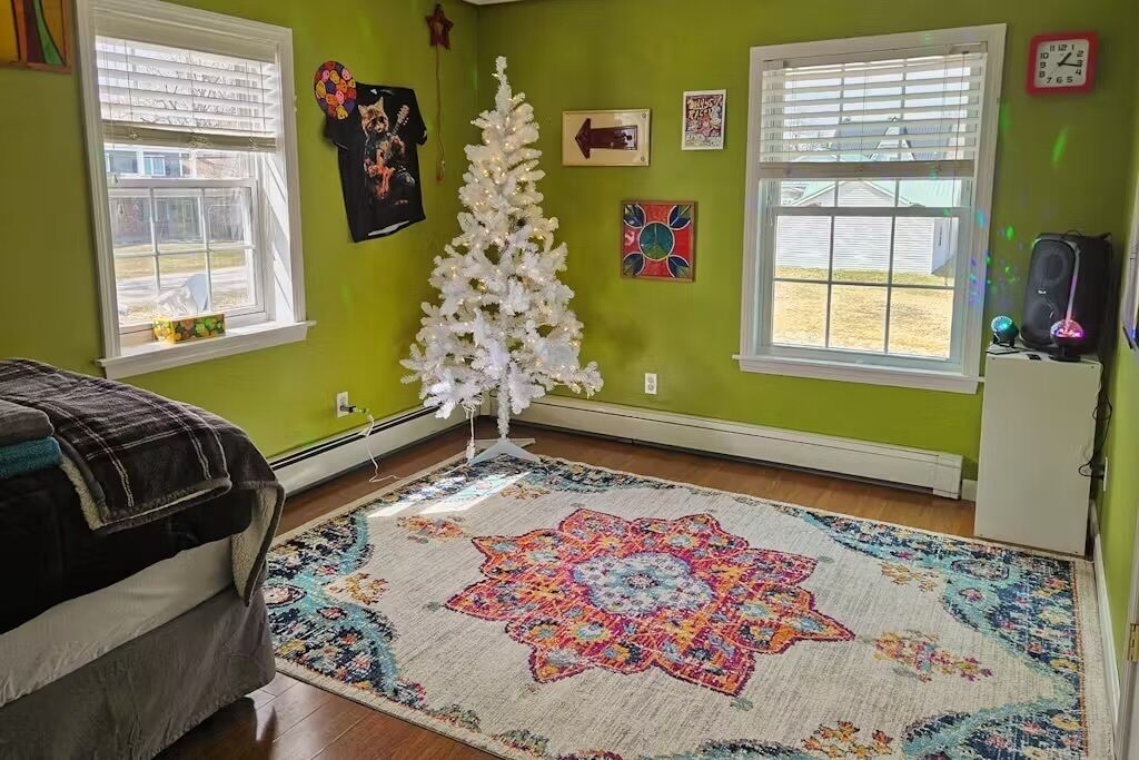 Une pièce avec un sapin de Noël blanc, un tapis coloré et un lit recouvert d’une couverture sombre.