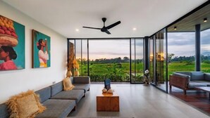 A modern living room with a large window, a ceiling fan, and a view of a green landscape. - Villa Eden by BaliSuperHost (Bali)
