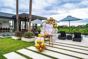 An outdoor wedding setup with a white arch and gold balloons, a pool, and lounge chairs. - Villa Eden by BaliSuperHost (Bali)