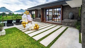 A modern house with a large glass door, a stone wall with a Buddha statue, and a grassy area with a balloon arrangement. - Villa Eden by BaliSuperHost (Bali)