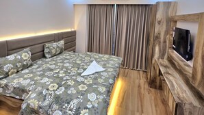 A modern bedroom with a bed, a ceiling fan, recessed lighting, an air conditioning unit, and a TV mounted on a wooden wall. - Anantya Inn (Greater Noida)