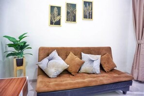 Un sofá marrón con tres cojines, un taburete de madera con una planta, y dos obras de arte con hojas enmarcadas en la pared.