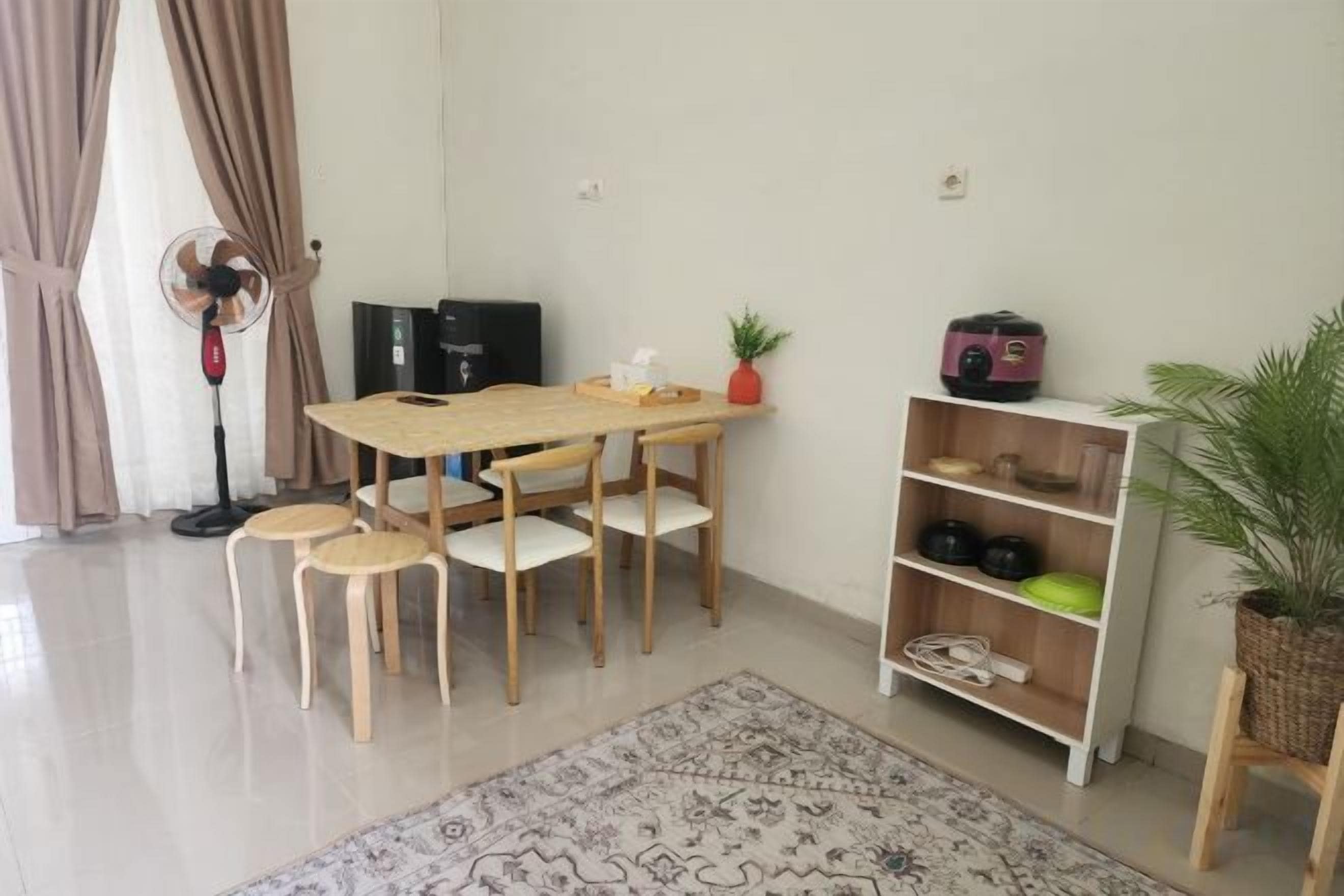 A dining area with a wooden table and chairs, a standing fan, a bookshelf, and a potted plant.
