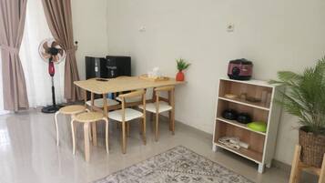 A dining area with a wooden table and chairs, a standing fan, a bookshelf, and a potted plant.