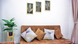 Een bruine bank met drie kussens, een klein houten krukje met een plant erop, en drie bladkunstwerken in lijsten aan de muur.