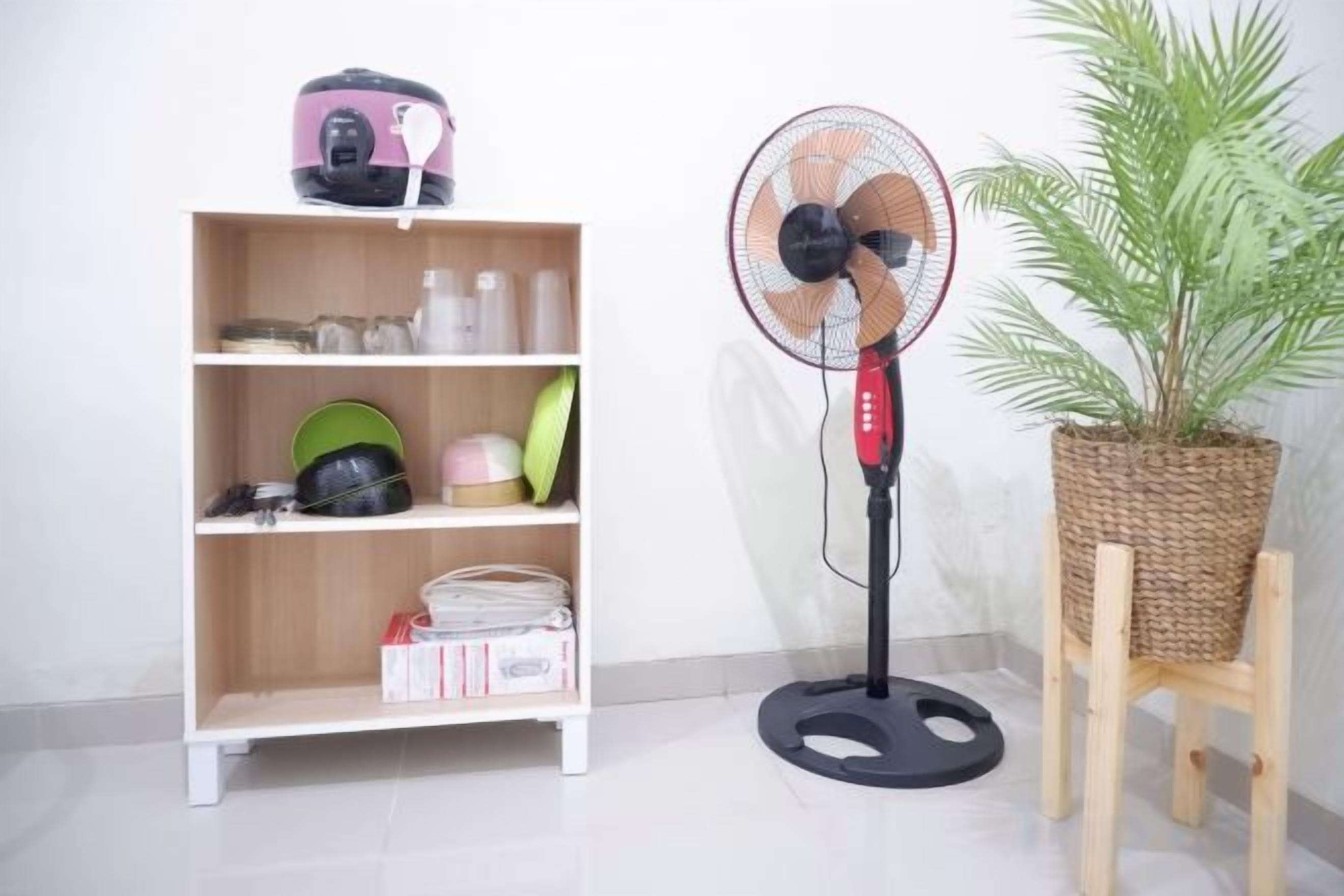 A room with a standing fan, a shelf with various items, and a potted plant.
