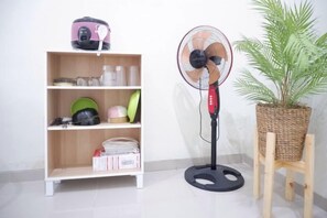 A room with a standing fan, a shelf with various items, and a potted plant. - Baturu Villa Kayana Regency D9 (Junrejo)