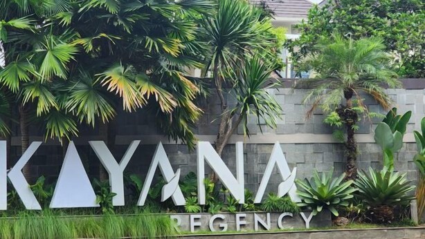 Een bordje voor 'Kayana Regency' met een stenen muur en tropische planten.