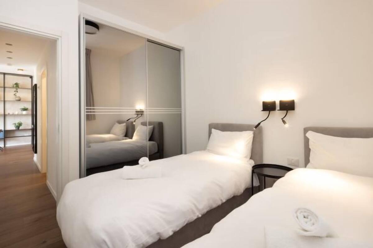 A modern hotel room with two beds, a sliding door closet, and wall-mounted lighting.