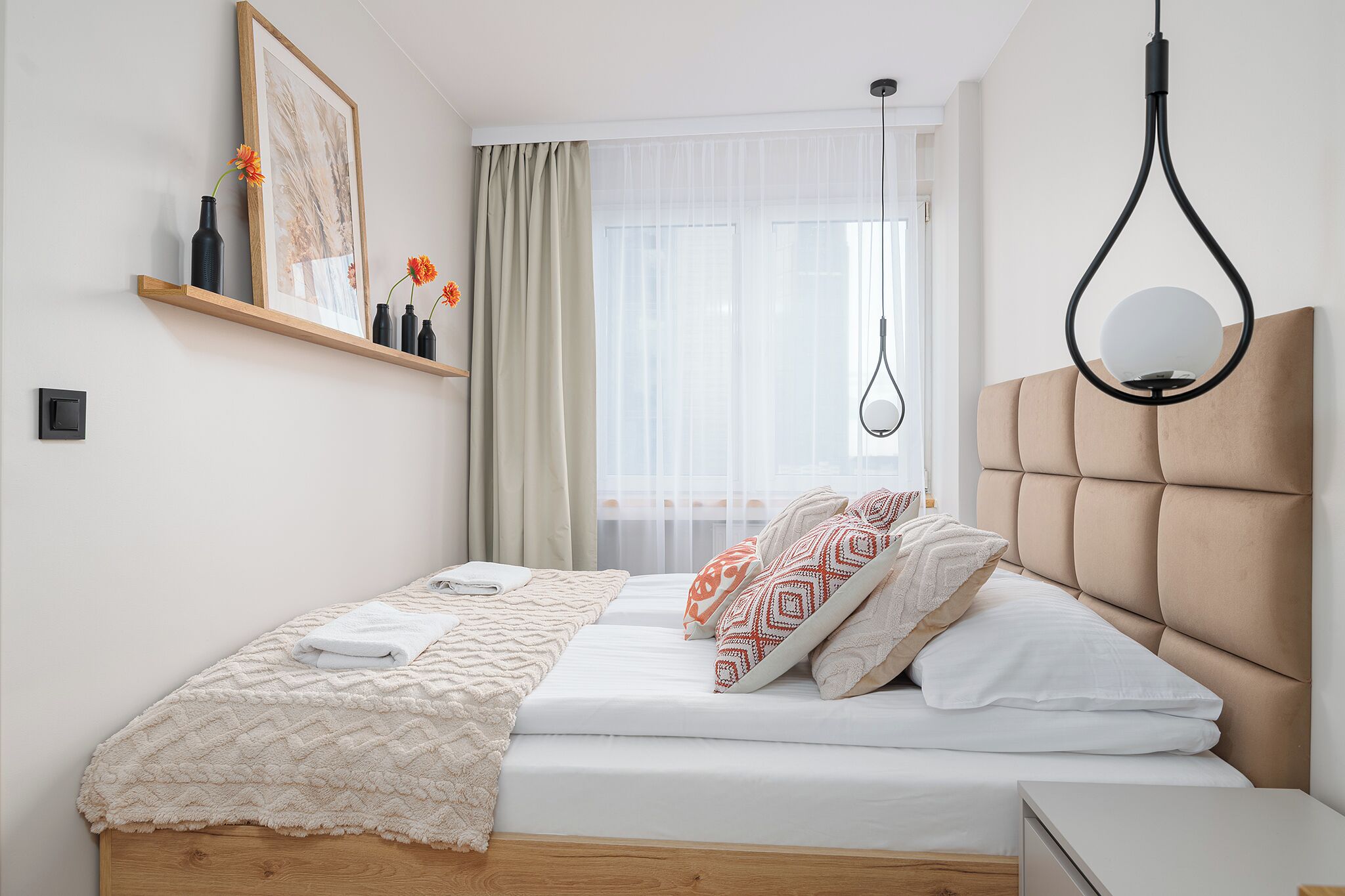 A neatly made bed with a white comforter and patterned pillows, a bedside table with a lamp, and a window with sheer curtains.