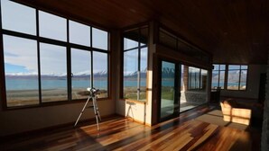 A spacious room with large windows offering a mountain view, wooden flooring, and a tripod in the corner. - Isolation Bay (Lake Tekapo)