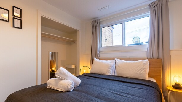A neatly made bed with a dark bedspread and white pillows, a bedside lamp, and a window with curtains. - 2 Bedrooms l  Sleeps 4 Adults l Kitchenette (Fernie)