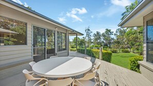 A covered outdoor patio with a round table and chairs, overlooking a well-maintained garden and a body of water.