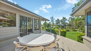 Un patio techado al aire libre con una mesa redonda y sillas, con vista a un jardín cuidado y una masa de agua.
