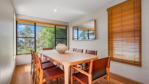 A dining area with a wooden table and chairs, large windows with blinds, and a brick wall.