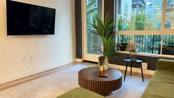A modern living room with a flat-screen TV, a round wooden coffee table, a green sofa, and a large window with a view of buildings and greenery.