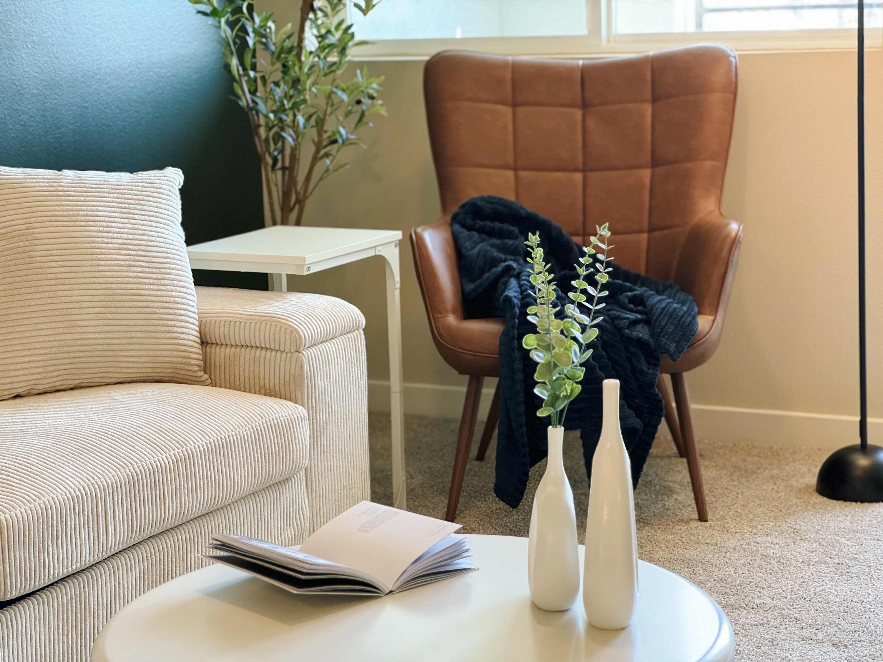 A living room with a beige sofa, a small white table, a brown armchair, and a plant in a white vase.