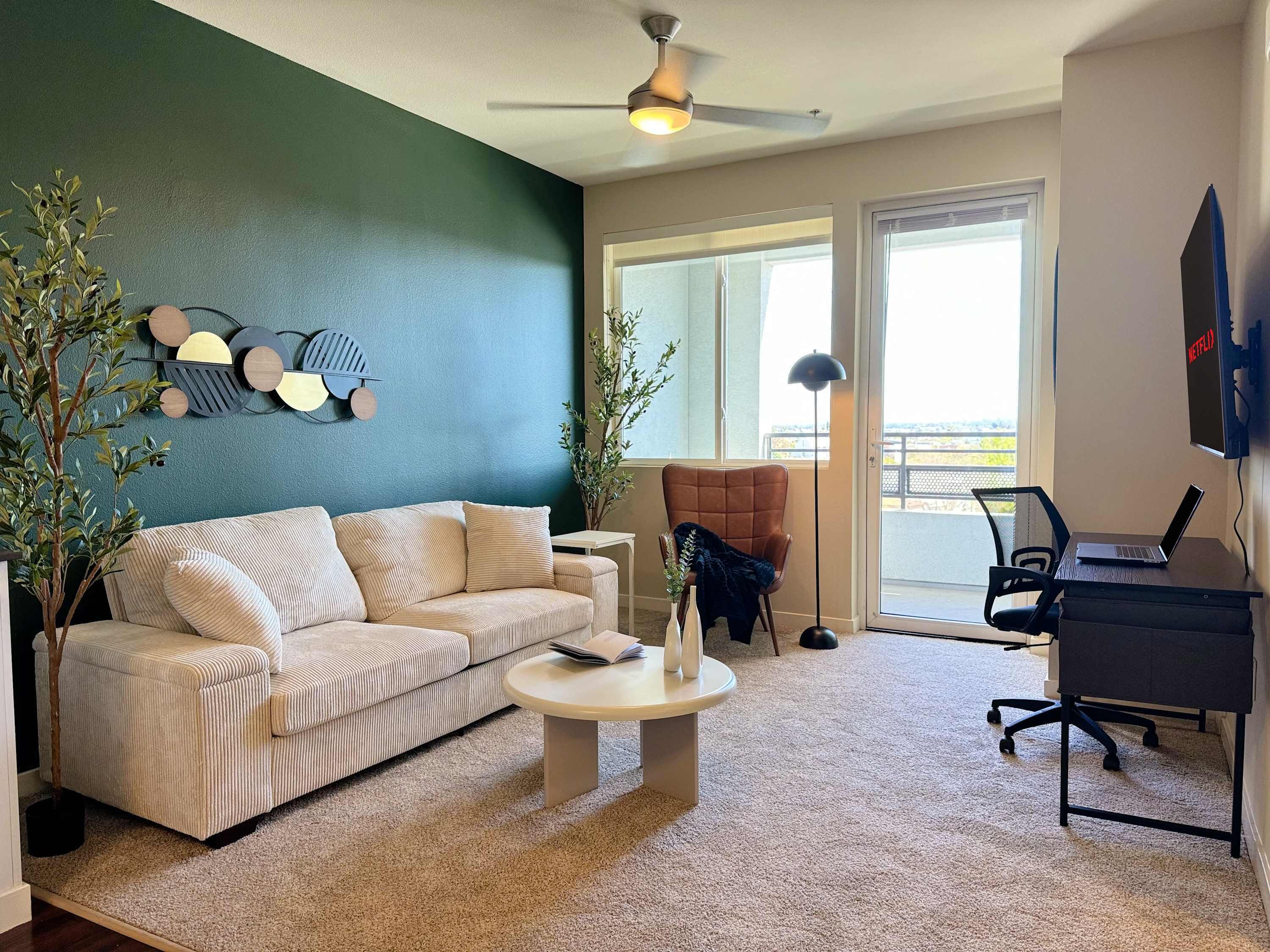 A living room with a beige sofa, a small round coffee table, a laptop on a desk, and a ceiling fan.