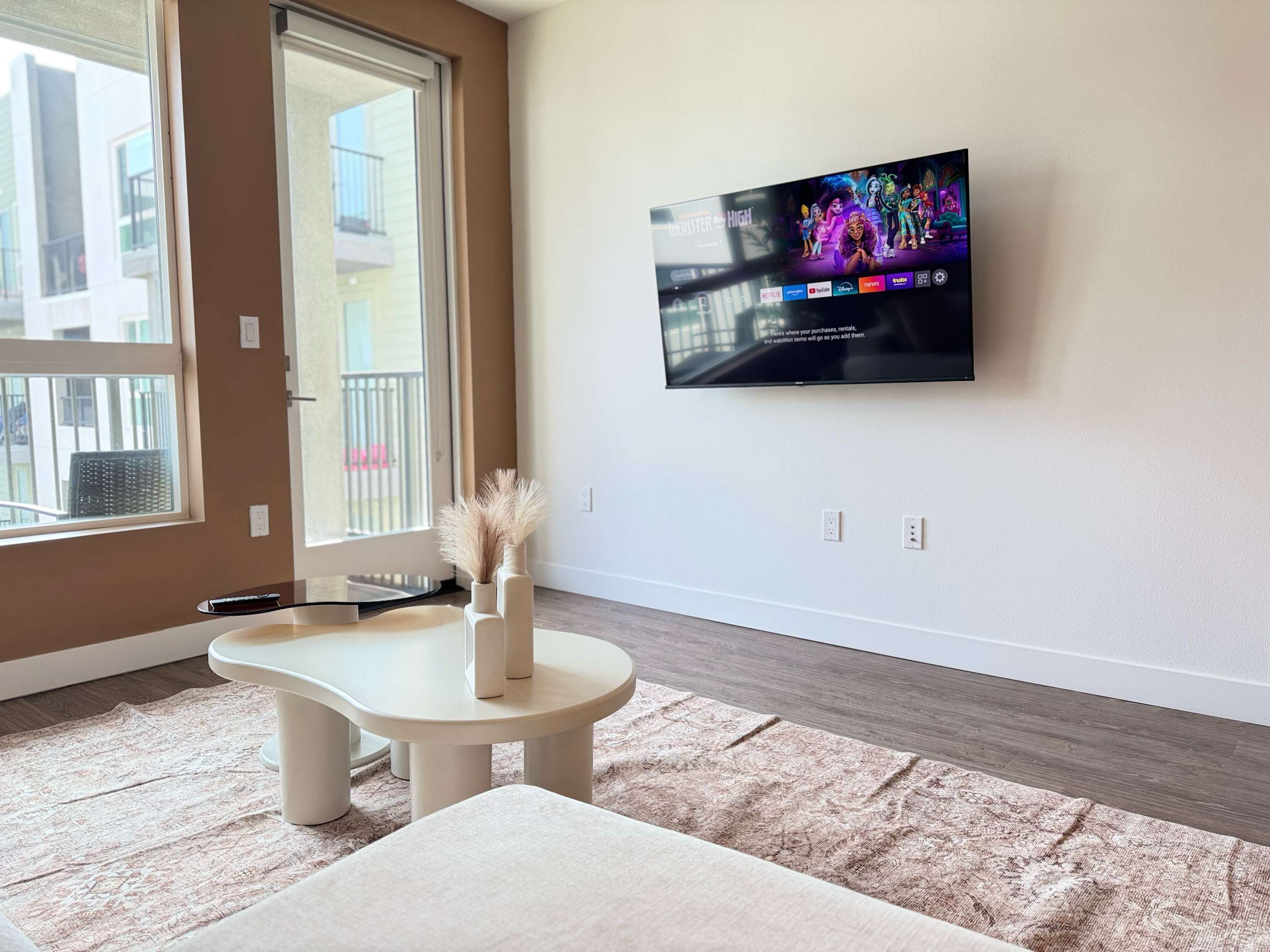 A modern living room with a flat-screen TV mounted on the wall, a small round table, a bench, and a decorative vase with pampas grass.