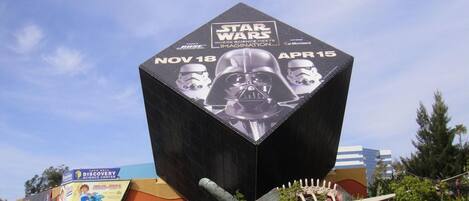 Eine große, Star Wars-bezogene Skulptur mit sechseckigem Sockel und gewölbter Spitze, befindet sich in einer Grünanlage, im Hintergrund ist ein Taco Bell Schild zu sehen.