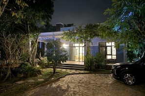 Un bâtiment avec une entrée pavée, une voiture garée devant et un jardin agrémenté d’arbres et d’arbustes.